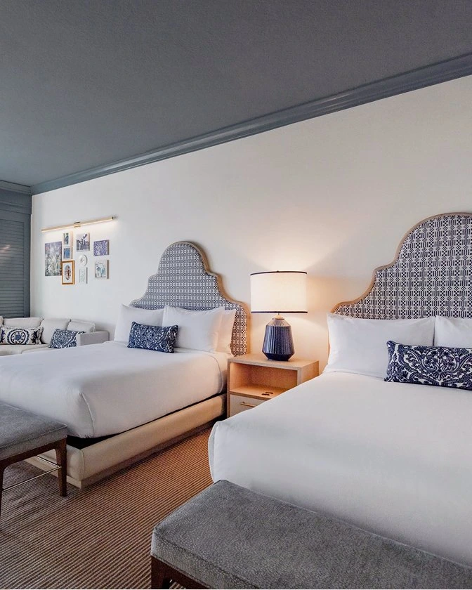 Two queen beds with blue and white patterned headboards in a bright Seagate Hotel deluxe room with a private balcony