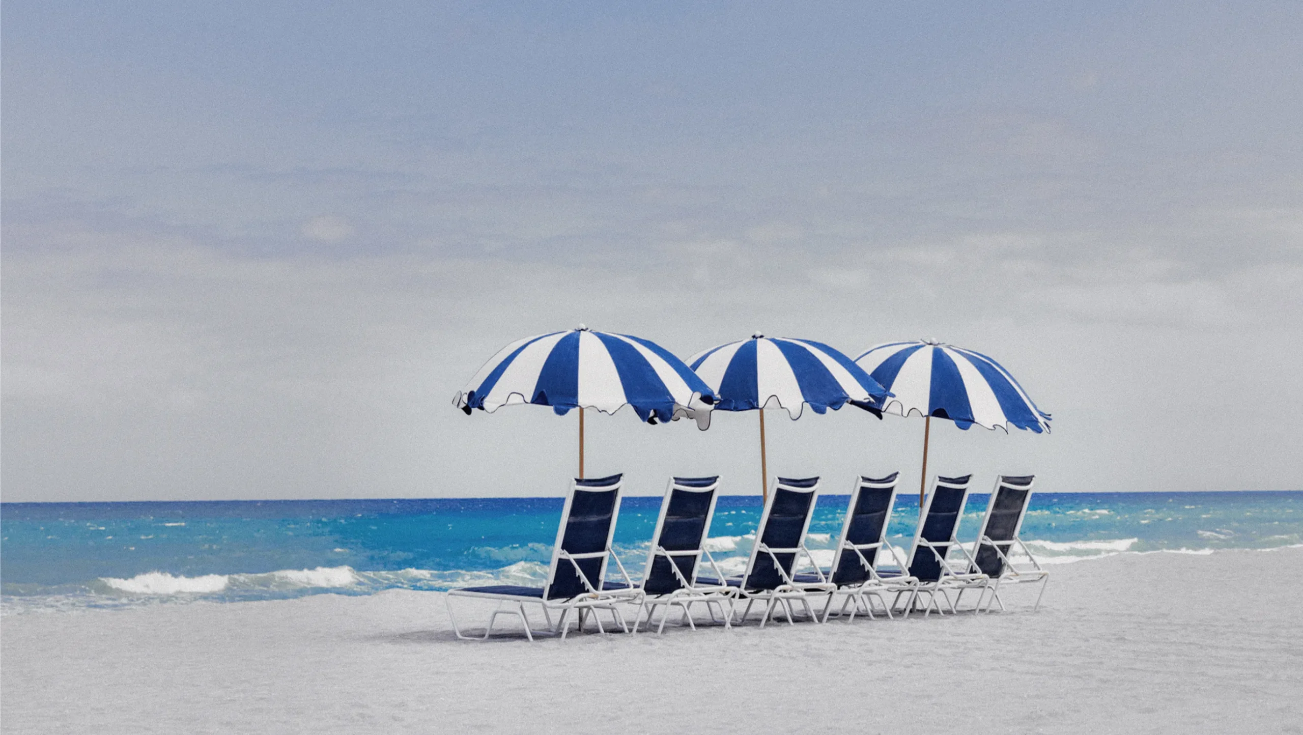 Six navy beach loungers under three blue and white striped umbrellas on the private member beach at The Seagate, ocean in background