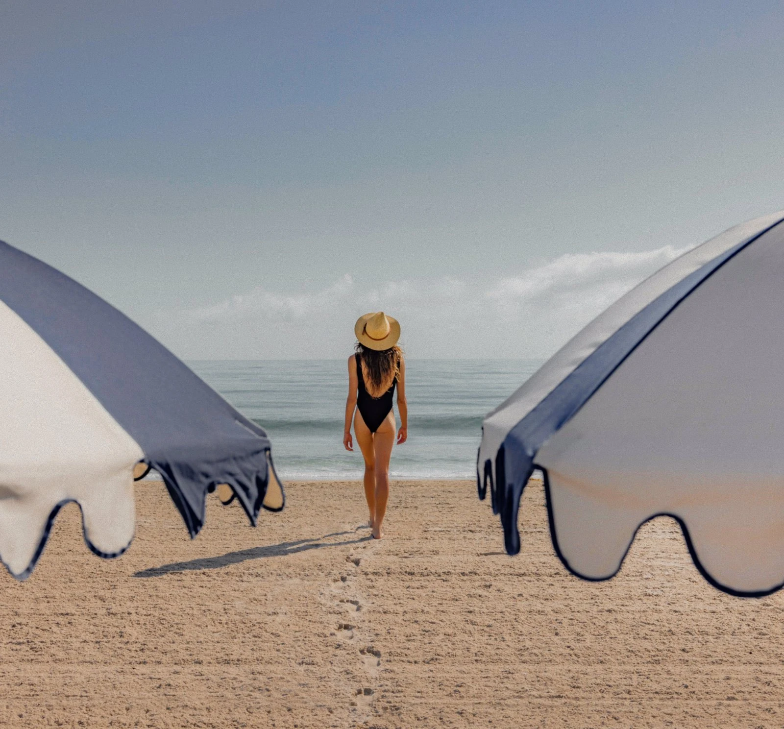 Woman in a swimsuit walking toward the ocean between two blue and white striped umbrellas on the private member beach at The Seagate